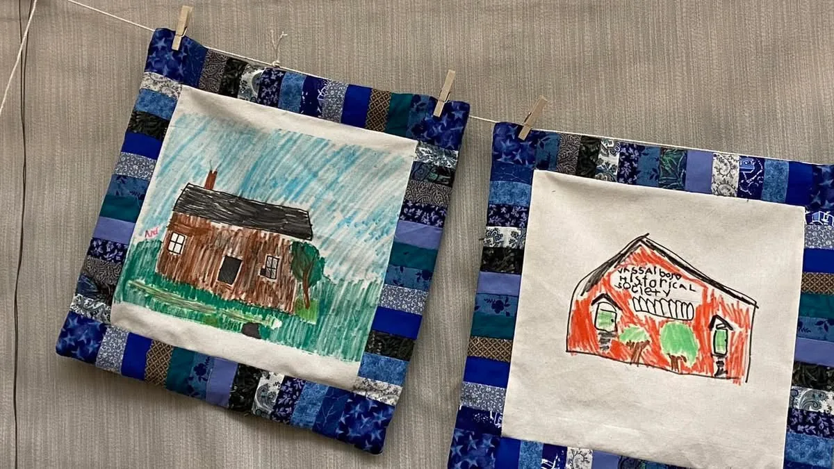 Two community quilt blocks pinned to a clothesline, showing a child's drawing of a house on the left, and a hand-drawn portrait of the Vassalboro Historical Society building on the right, both bordered in strip-pieced blue patchwork