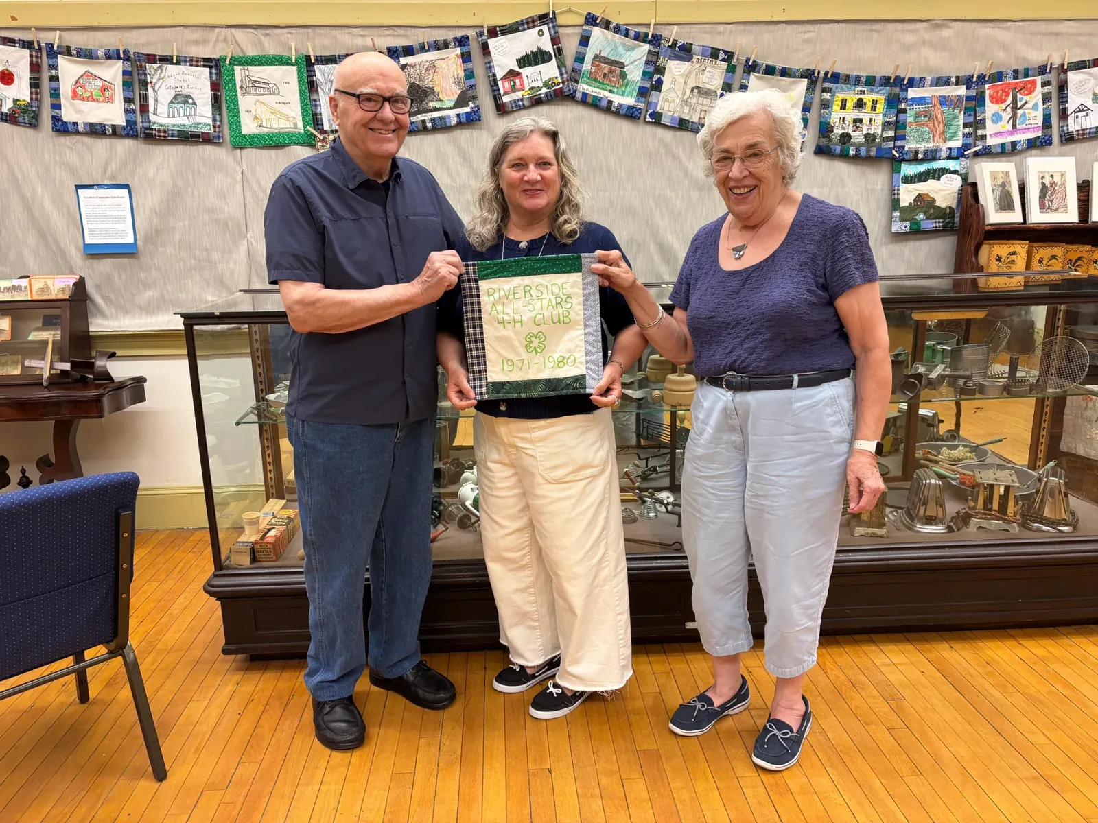 Three volunteers stand in front of a clothesline strung with dozens of hand-decorated community quilt blocks at the Vassalboro Historical Society, holding a green-bordered Riverside All-Stars 4-H Club 1971-1980 block