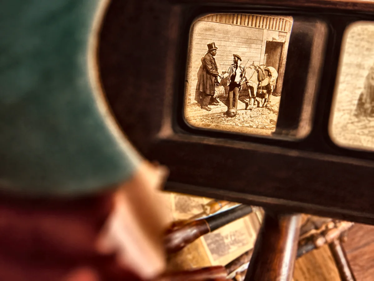 View through a stereoscope viewer showing a sepia card of a top hatted man and a groom with horses outside a stable