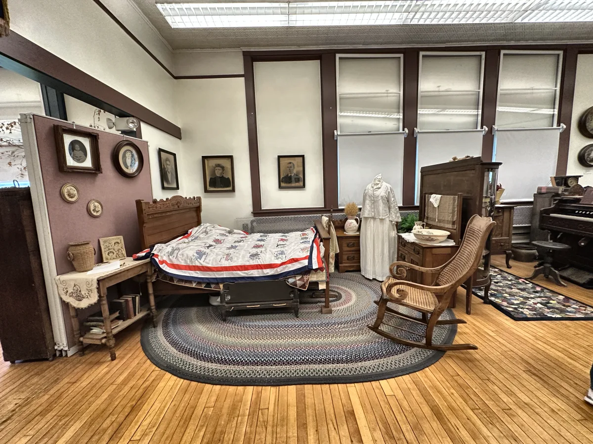 Period bedroom with a signature quilt on a carved bed, washstand, braided rug and rocking chair