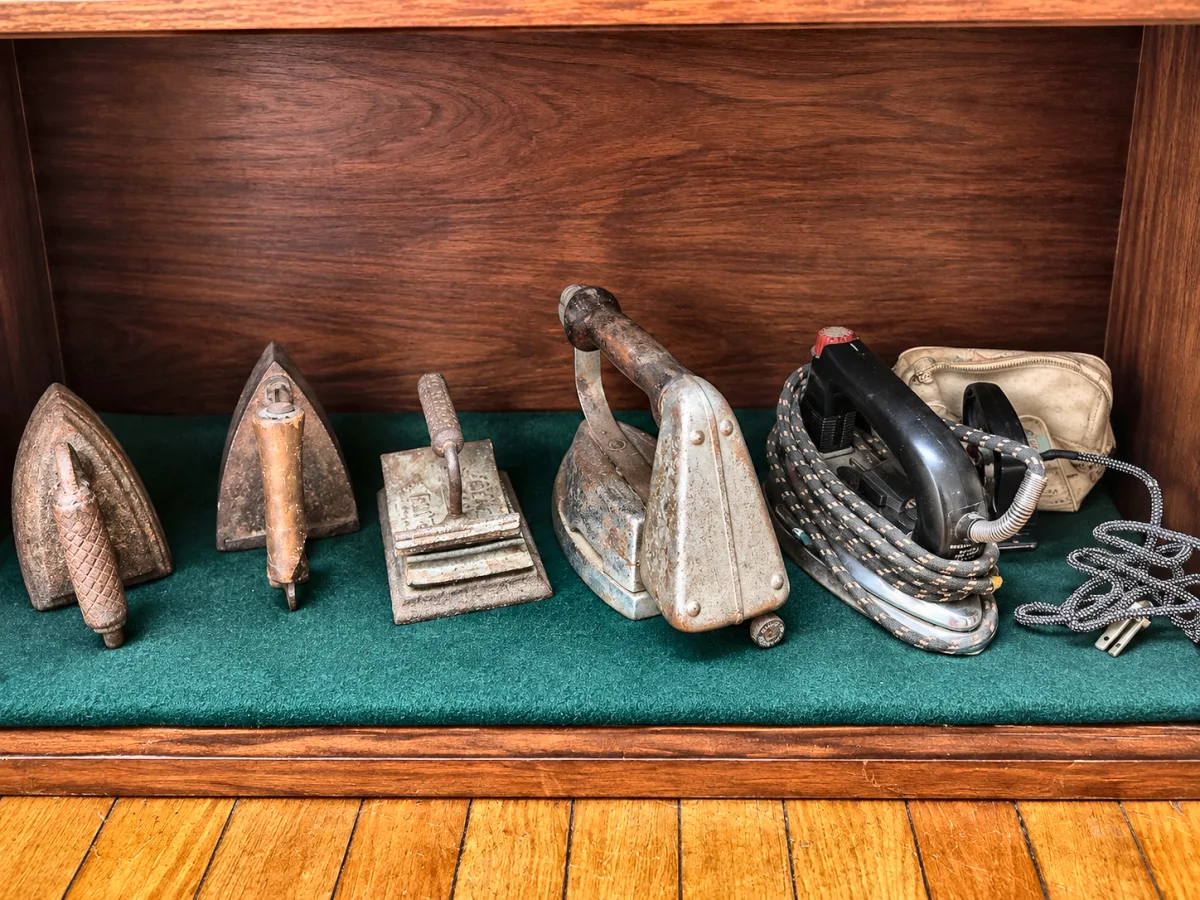 Row of five irons from cast iron flatirons to an early electric iron with a braided cord