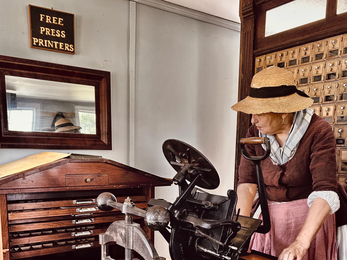 Curator Susan Reidy in period dress working the platen press beneath the Free Press Printers sign
