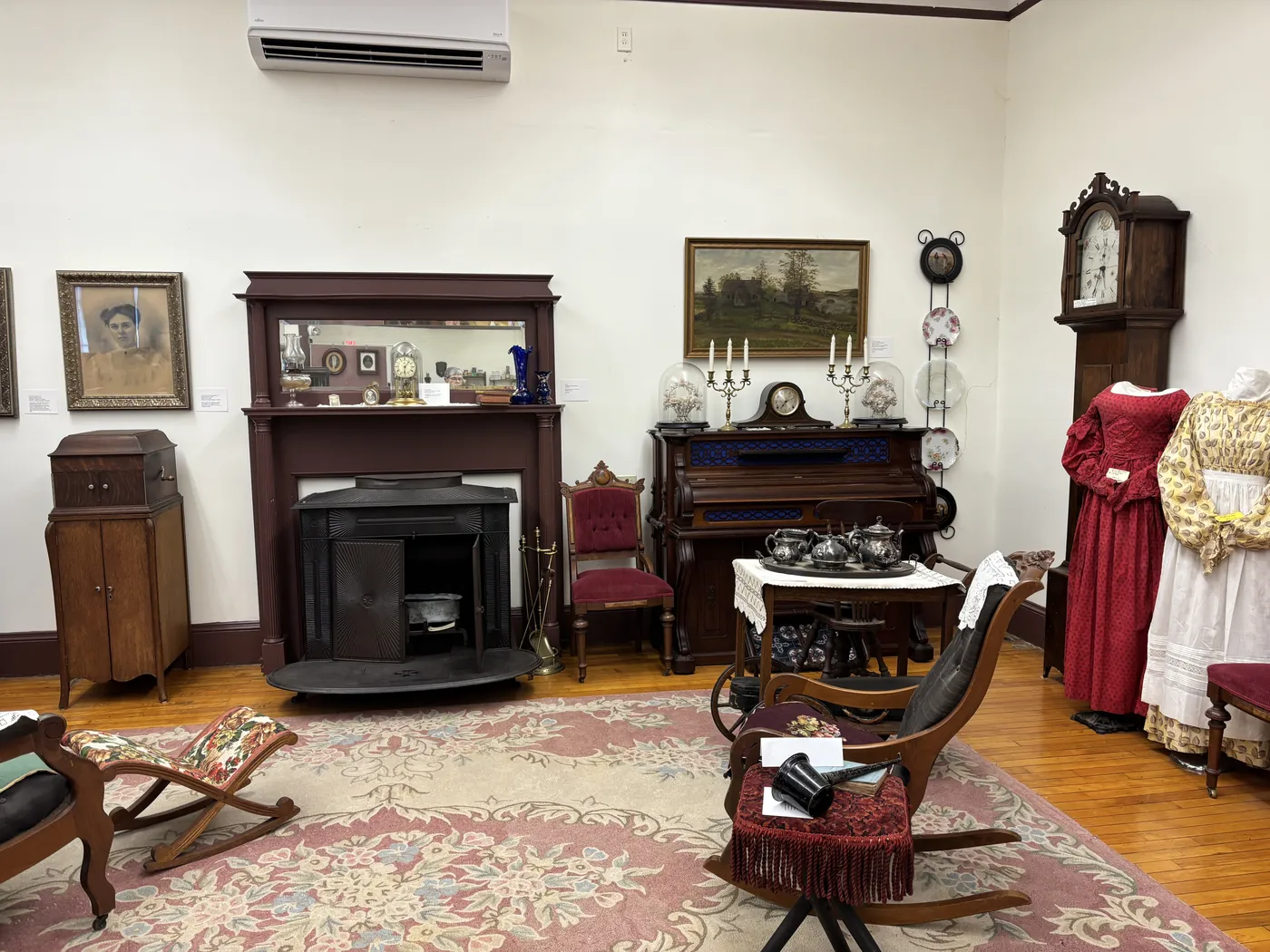 A Vassalboro parlor setting with a period fireplace, parlor organ with candelabras and silver tea service, Victorian red dress, grandfather clock, and floral rug