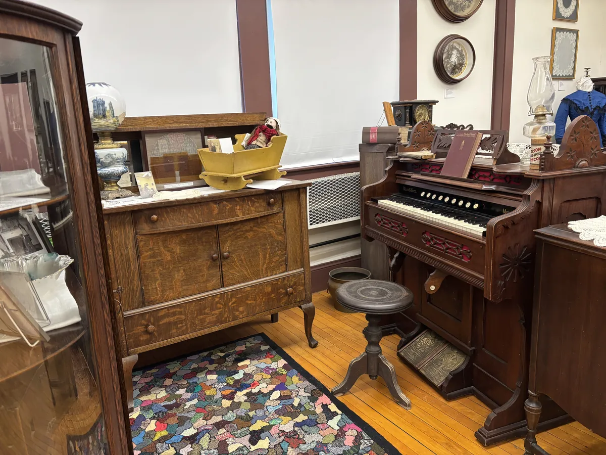 Close view of a Wilcox & White pump organ with red felt detail, an antique doll cradle on a nearby dresser, and a hand-hooked rug on the wood floor