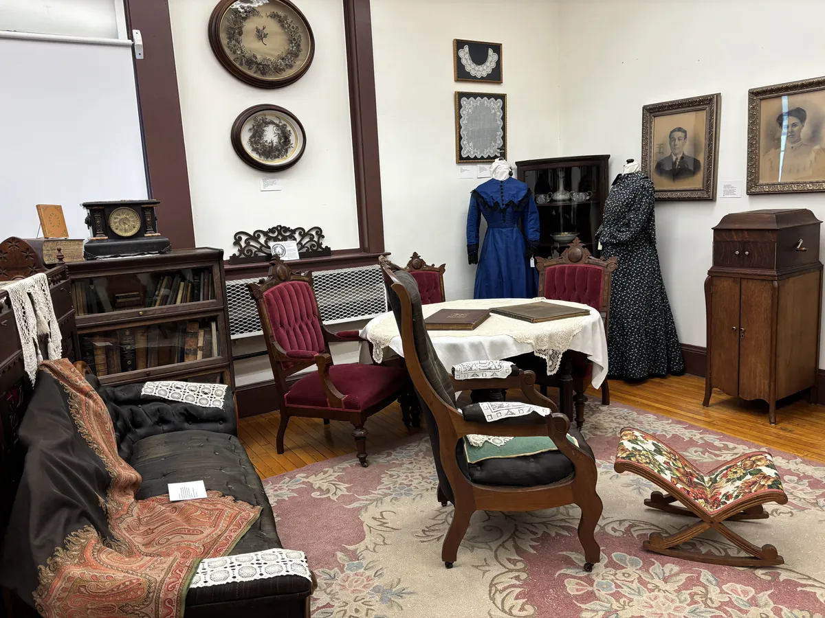 Victorian sitting room with a black chaise, lace-topped table with a family Bible, two mannequins in period dresses, and framed portraits of a Victorian couple on the wall