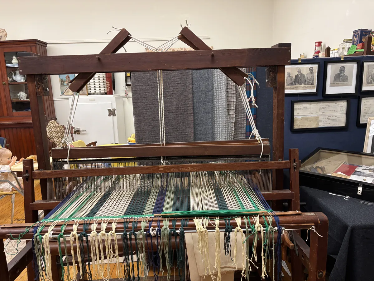 A large working loom in the museum, strung with green and cream warp threads and showing woven fabric samples in shades of blue and grey