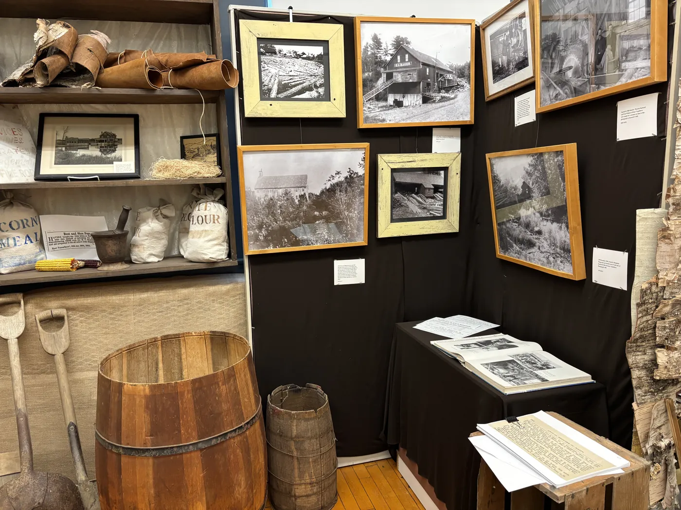 A wall of framed historical photographs of Vassalboro mills and logging operations, alongside period grain and flour sacks, a mortar and pestle, and coopered wooden barrels