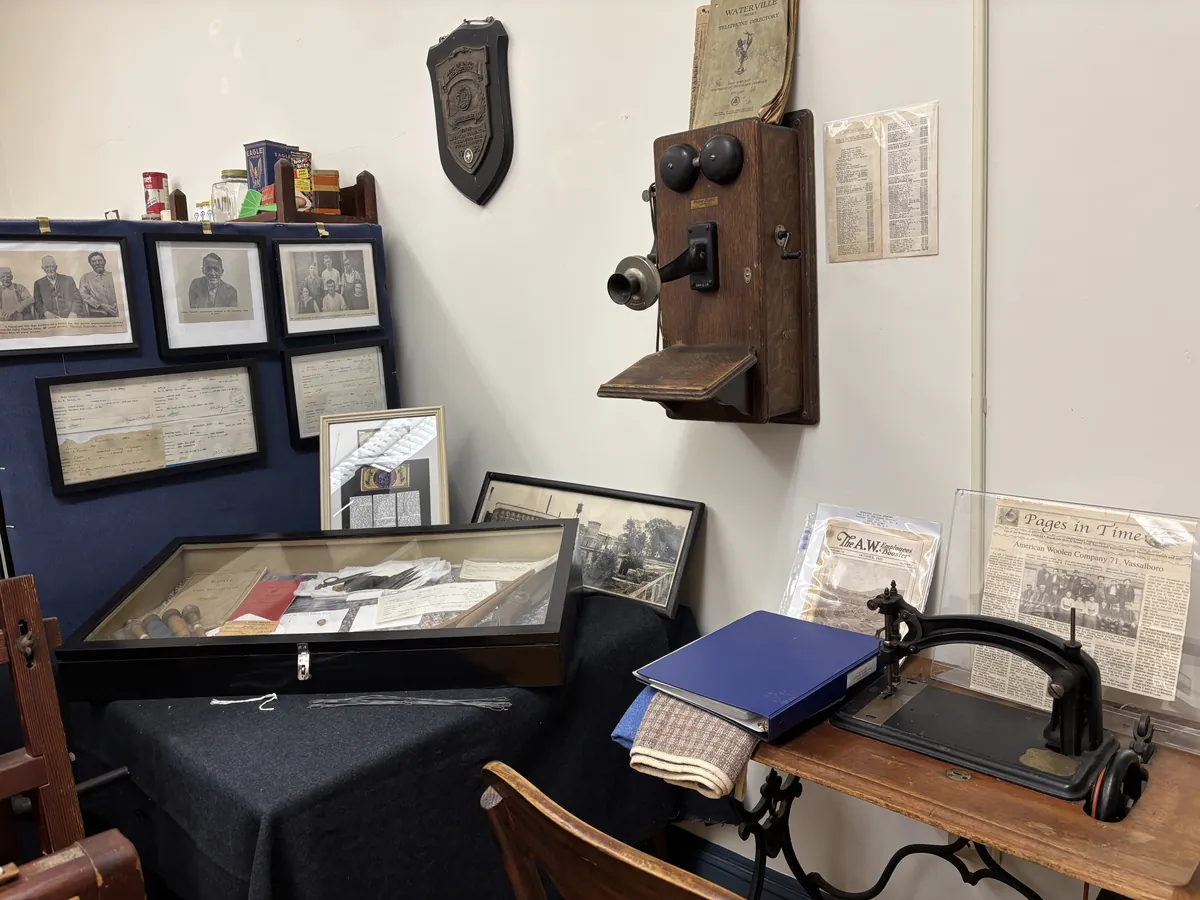 North Vassalboro Woolen Mill display: portraits of long-serving mill workers, framed tax and mill records, an oak wall telephone, a wool sample case, and a leather sewing machine