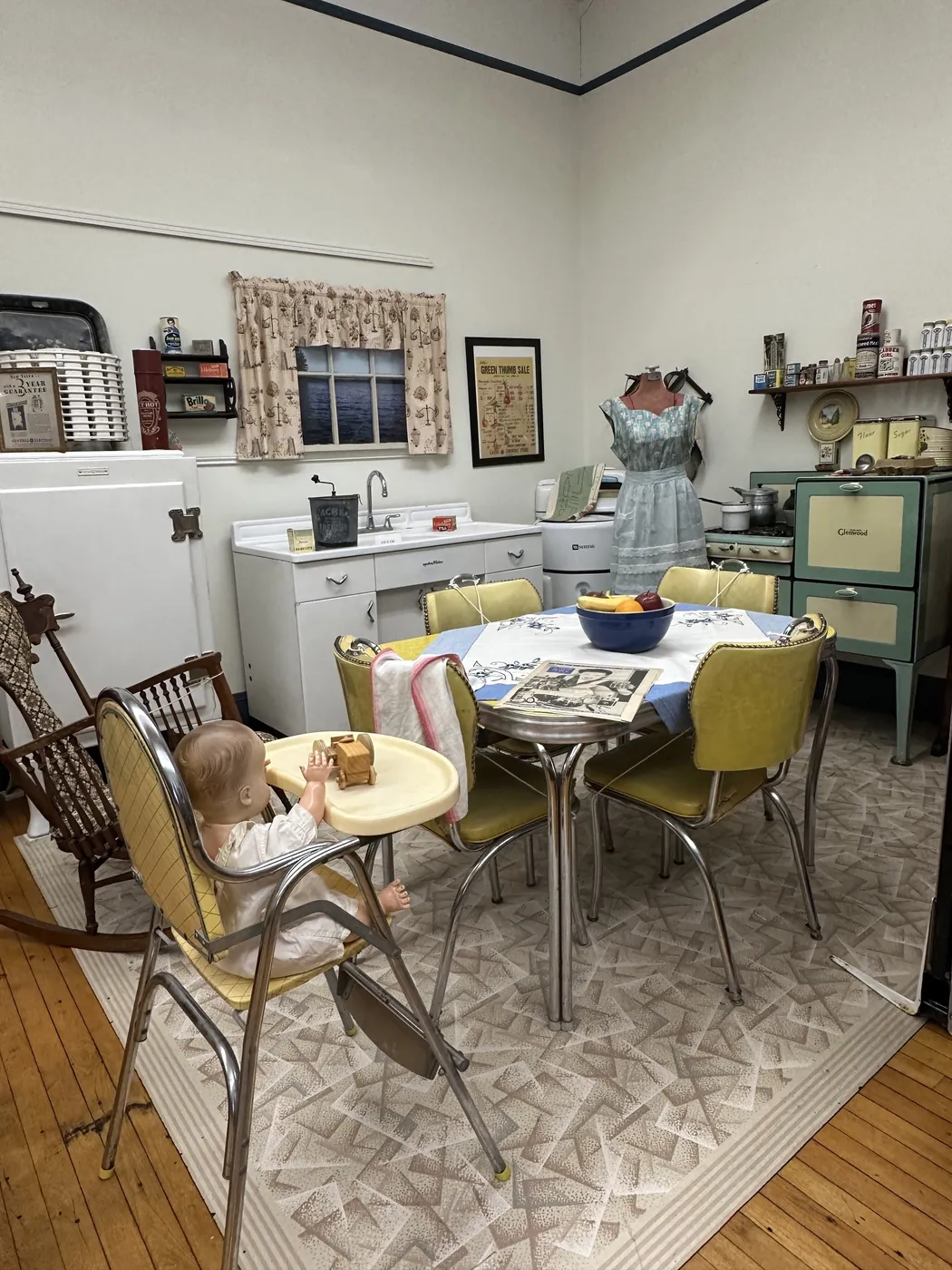 A staged mid-century kitchen at the museum with a Glenwood range, white Maytag icebox, chrome dinette set with yellow chairs, and a baby doll in a period high chair