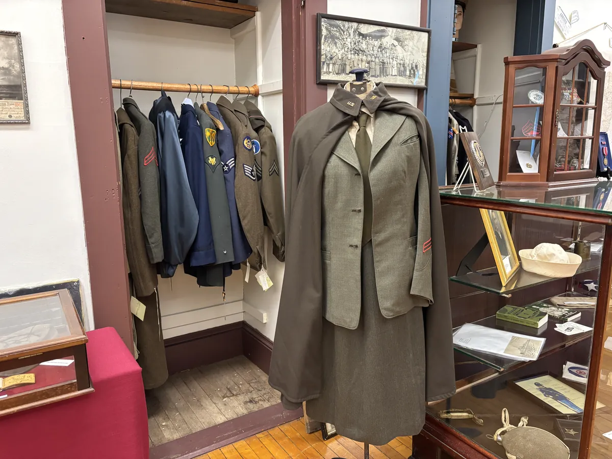 A rack of Army and Air Force uniforms next to a mannequin dressed in a WWII-era WAC uniform with cape, at the Vassalboro Historical Society