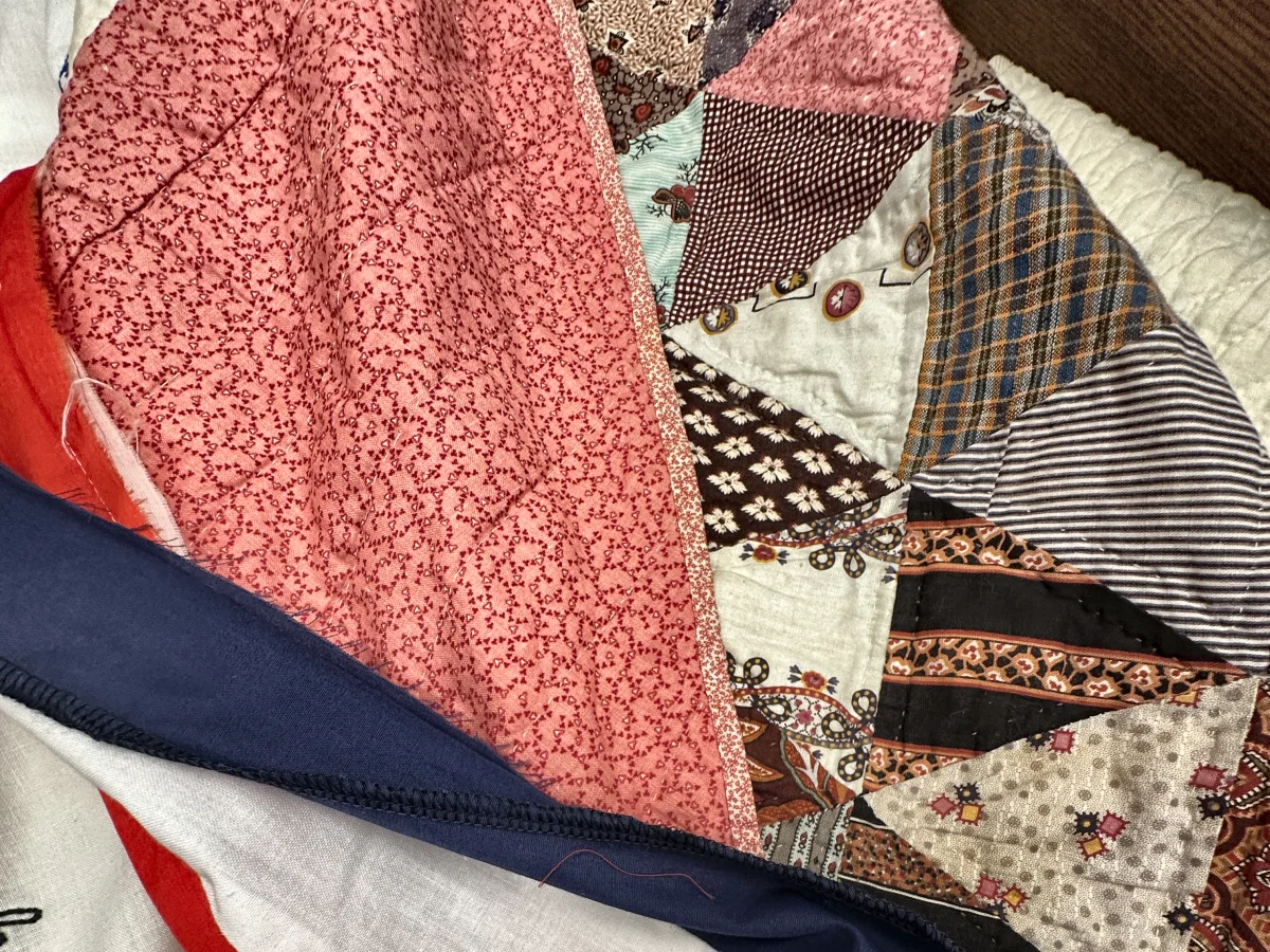 Three quilts folded together: a crazy quilt of pieced silks and cottons, a pink printed quilt, and a red, white and blue embroidered coverlet