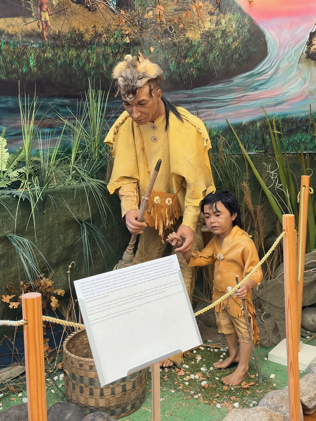 Native American father and son mannequins in leather clothing, positioned at a fishing scene with a painted background of a stream and grasslands — part of the Remembered Vision diorama at the Vassalboro Historical Society