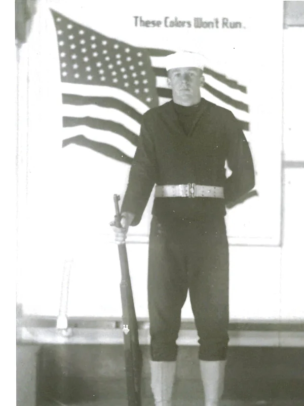 Gerald Robbins in his U.S. Navy uniform, 1943, standing at attention before an American flag with the caption 'These Colors Won't Run'